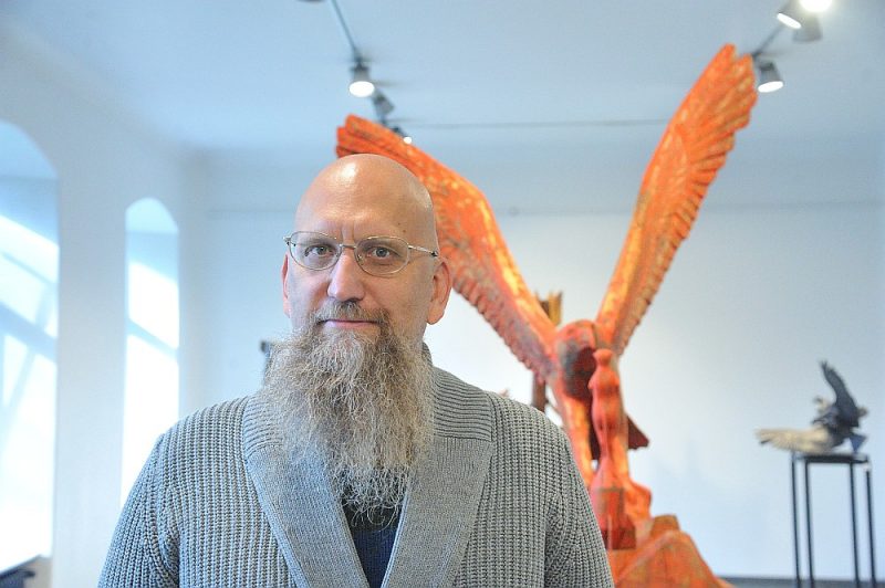 Aigars Bikše: “Arī mūsu modernajā sabiedrībā joprojām ir daudz arhaisma elementu.” Fonā: darbs “Putns aprauga surikatu”. Āfrikas dienvidi. Koks.